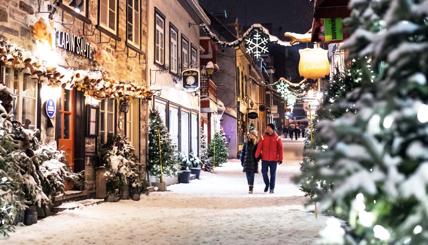A couple takes a walk in the evening during the holiday season on rue du Petit-Champlain, covered with snow and decorated with illuminated trees.