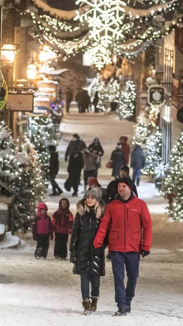 Plusieurs personnes se promènent en soirée sur la rue du Petit-Champlain enneigée et décorée de nombreux sapins illuminés. 