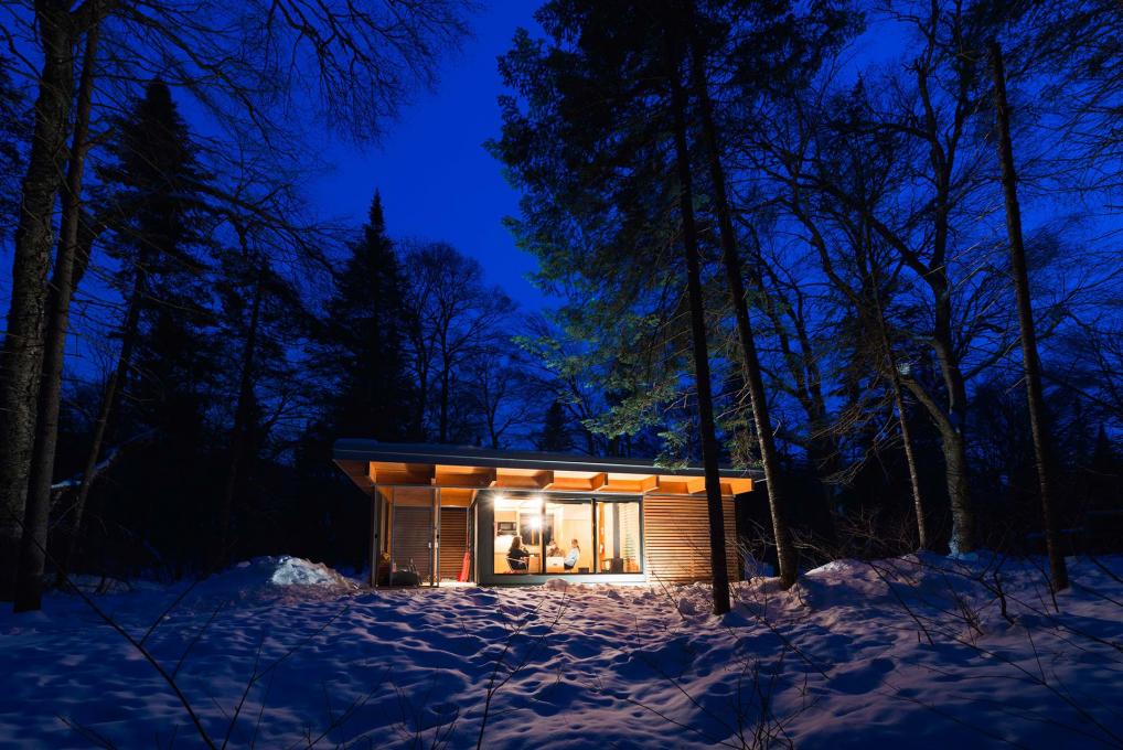 Chalet EXP illuminé en soirée, au coeur de la forêt dans le parc national de la Jacques-Cartier, en hiver.