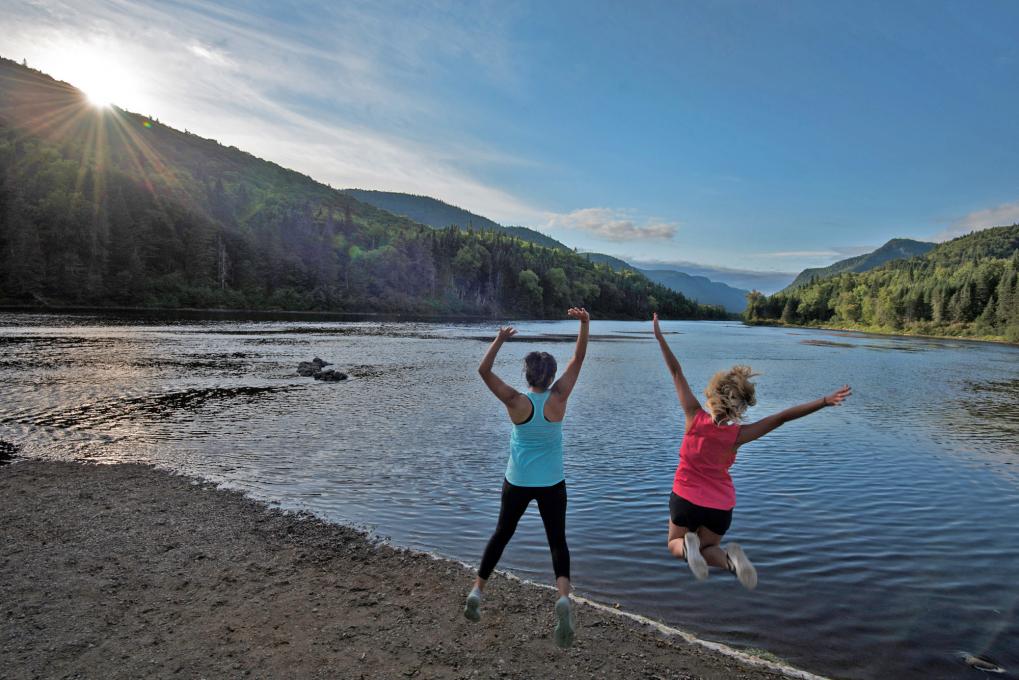 Two girls jump with joy in front of the Jacques-Cartier Valley, in Jacques-Cartier National Park in summer.