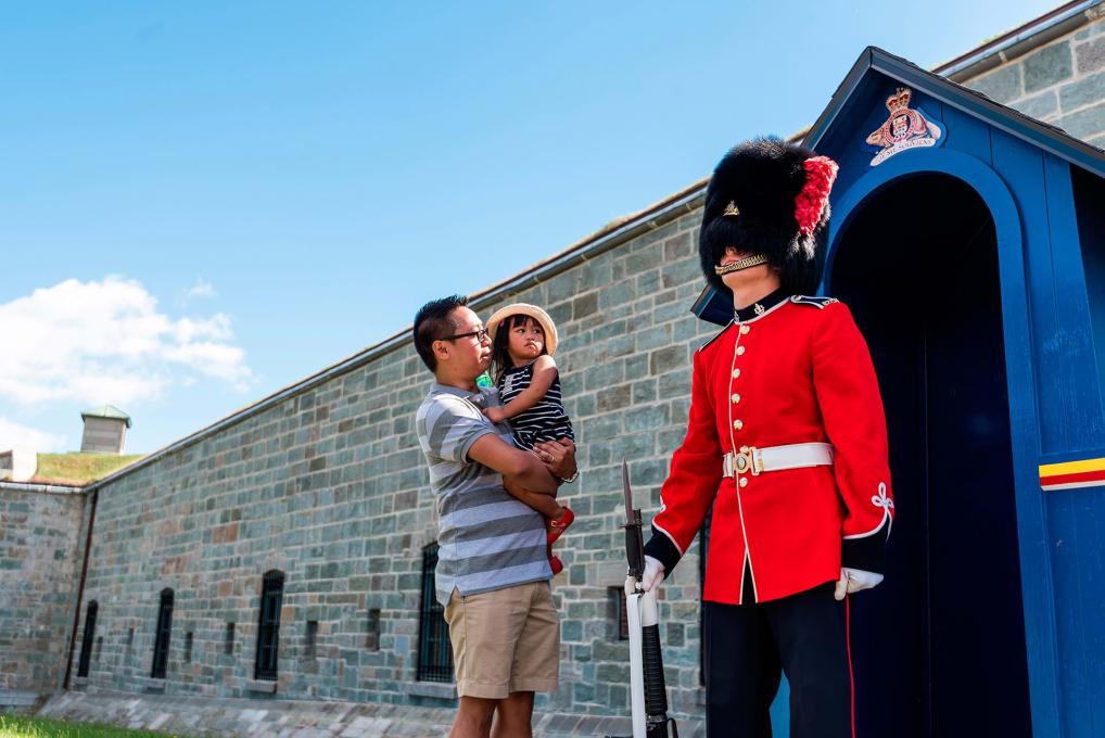 Un père et sa fillette regardent un garde vêtu de l'uniforme traditionnel devant la porte d'entrée de la Citadelle de Québec.
