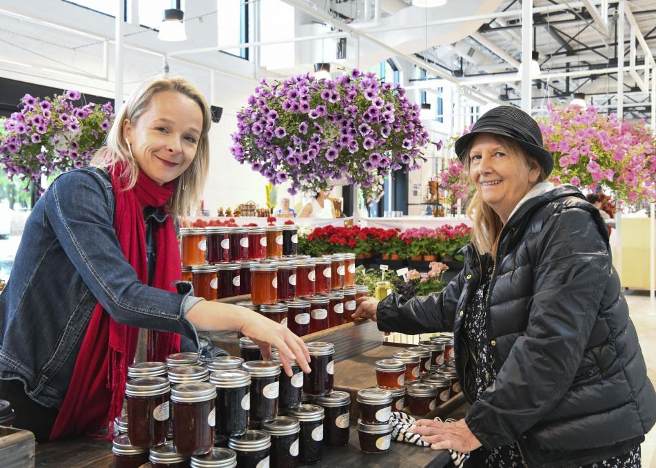 Two women near a kiosk of jars of jam and flowers at the Grand Marché of Québec.