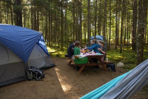A family eats a meal on a picnic table in the forest, at the Vallée Bras-du-Nord campsite.