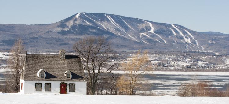 Heritage house on Ile d'Orléans in winter, with a view of the St. Lawrence River and the snow-covered ski slopes of Mont-Sainte-Anne in the background.