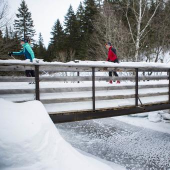 Deux personnes en ski de fond au Mont-Sainte-Anne, traversant un pont au dessus d'une rivière.