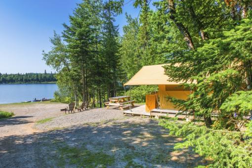 Vue extérieure d'un hébergement prêt-à-camper près d'un lac dans la Réserve faunique des Laurentide, avec table à pique-nique, chaises et chaloupes.