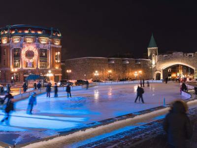 People on the outdoor ice rink at Place D'Youville, near the Capitol, the fortifications and the illuminated Saint-Jean gate.