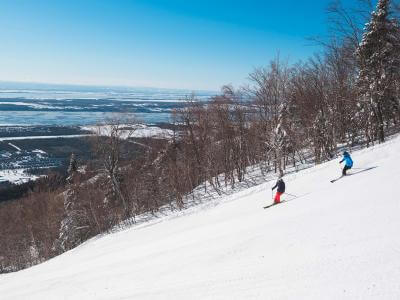 Two skiers go downhill skiing at Mont-Sainte-Anne station with a view of the St. Lawrence River.