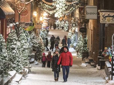 People walk in the evening on rue du Petit-Champlain, covered with snow and decorated with many illuminated trees.
