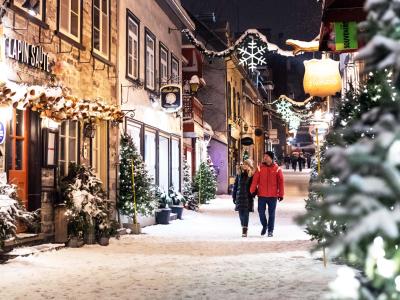 A couple takes a walk in the evening during the holiday season on rue du Petit-Champlain, covered with snow and decorated with illuminated trees.