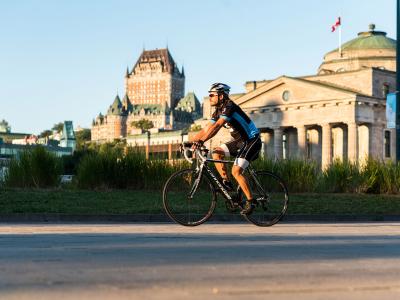 Un cycliste à vélo roule sur une piste cyclable dans le Vieux-Port de Québec avec la Château Frontenac en arrière-plan.