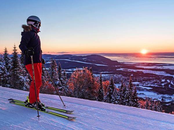 Une skieuse en ski alpin prend une pause et observe le coucher du soleil au sommet d'une montagne au Mont-Sainte-Anne.