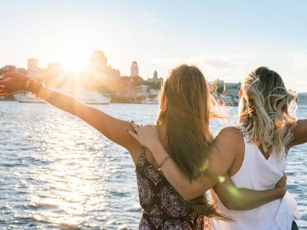 Two girls stand arm in arm on the Québec-Lévis ferry, in front of a panorama of Old Québec in summer.