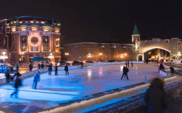 Des gens sur la patinoire extérieure de Place D'Youville, près du Capitole, des fortifications et de la porte Saint-Jean illuminée.