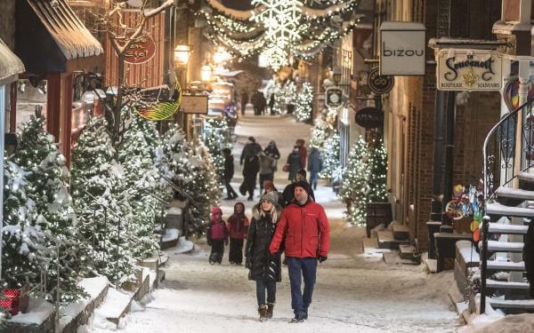  Several people walk in the evening on rue du Petit-Champlain, covered with snow and decorated with many illuminated trees.