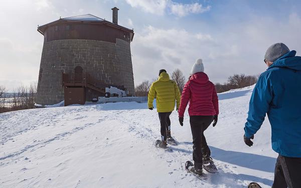 Three friends are snowshoeing on the snow-capped Plains of Abraham and heading towards the historic Martello Tower.