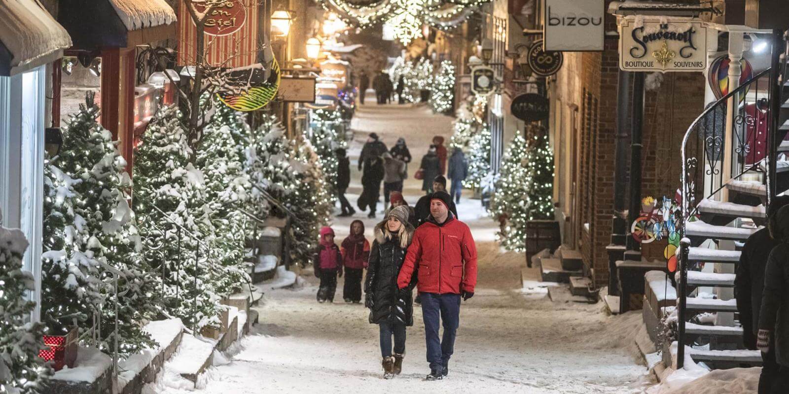 Plusieurs personnes se promènent en soirée sur la rue du Petit-Champlain enneigée et décorée de nombreux sapins illuminés. 