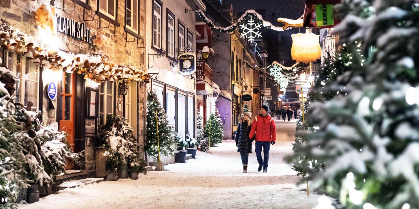A couple takes a walk in the evening during the holiday season on rue du Petit-Champlain, covered with snow and decorated with illuminated trees.