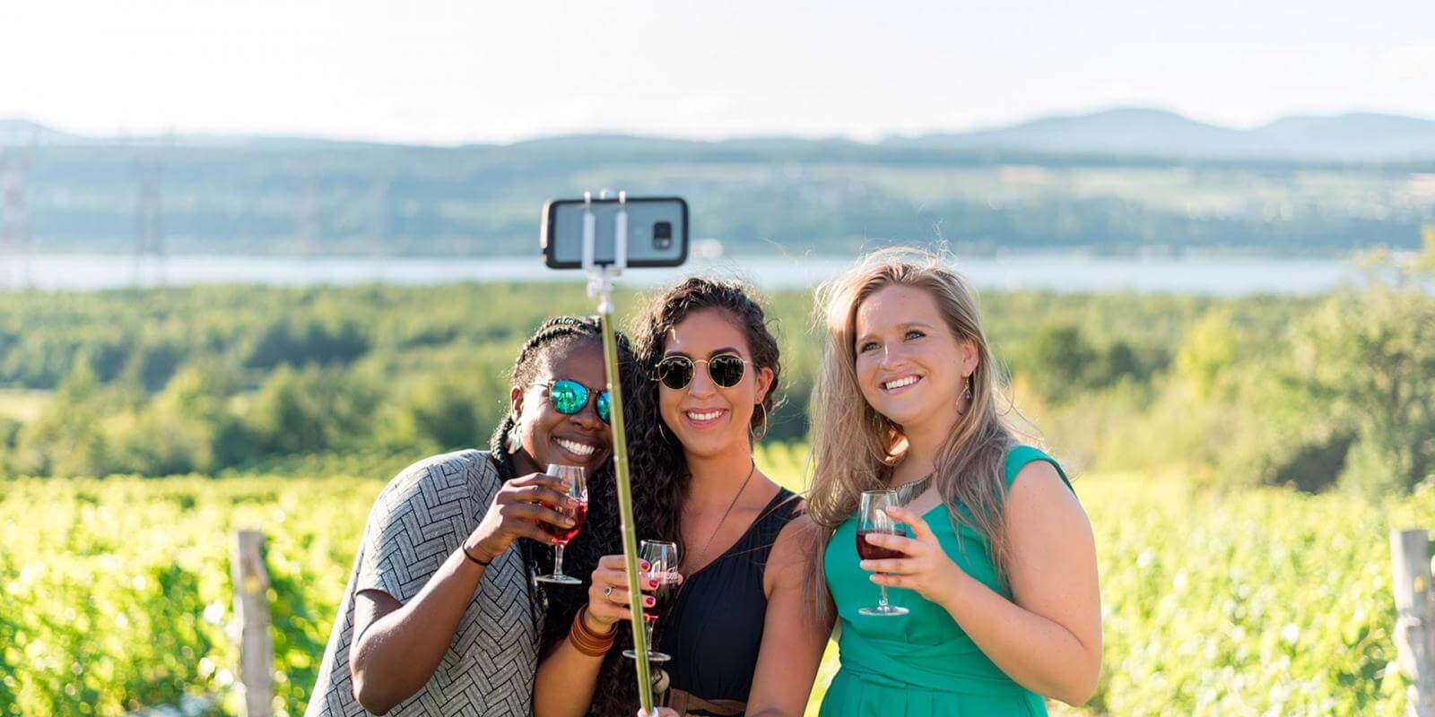 Three smiling girls take pictures of themselves and have a drink in the middle of a vineyard in Ile d'Orleans, in summer.