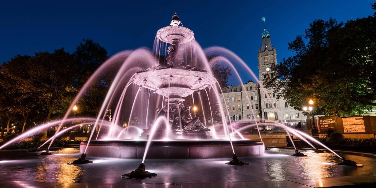 View of the Tourny Fountain illuminated in the evening and the Parliament Building in the background.