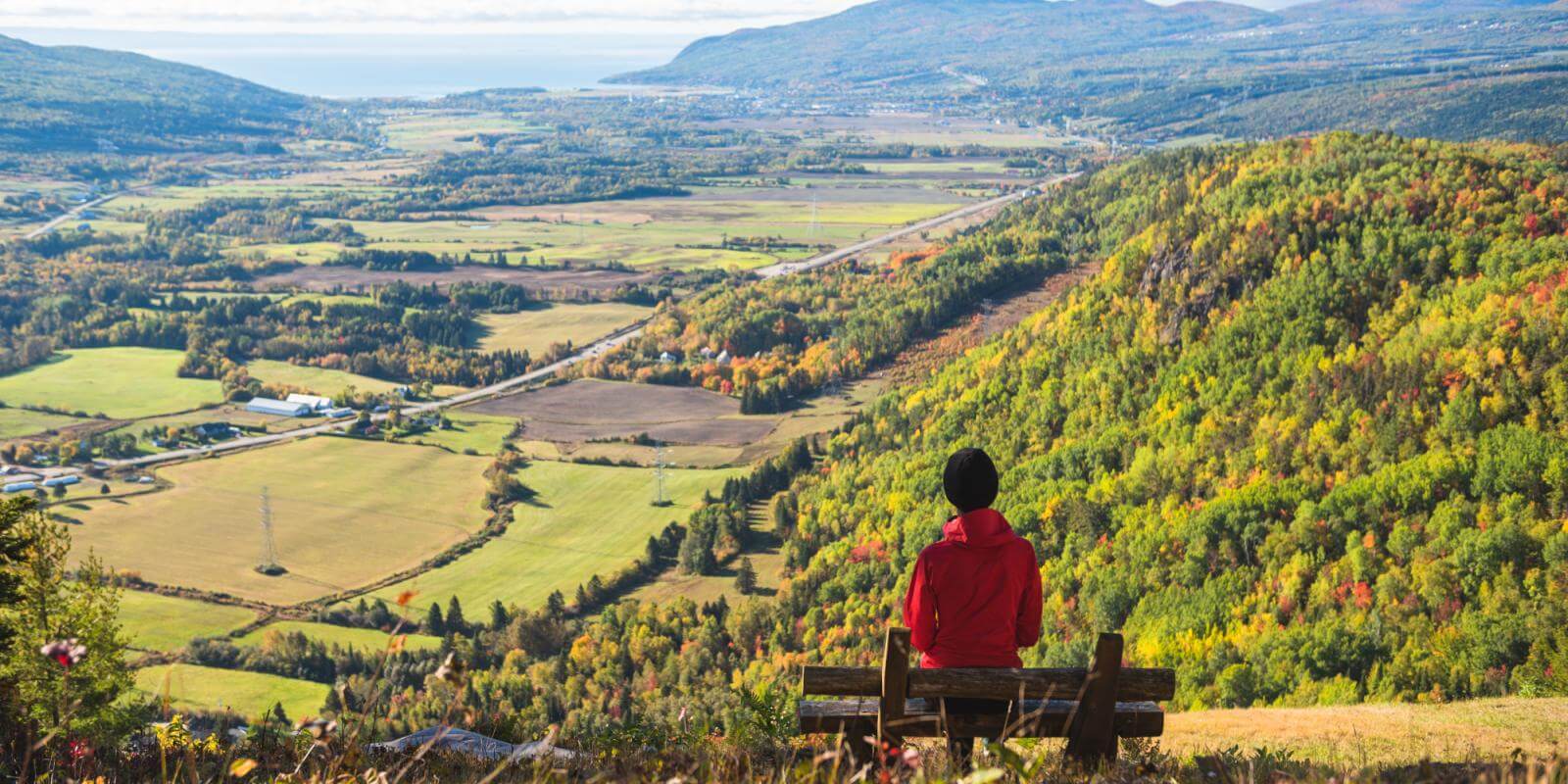 Une femme assise sur un banc au sommet d'une montagne observe le panorama et les couleurs d'automne dans la région de Charlevoix.