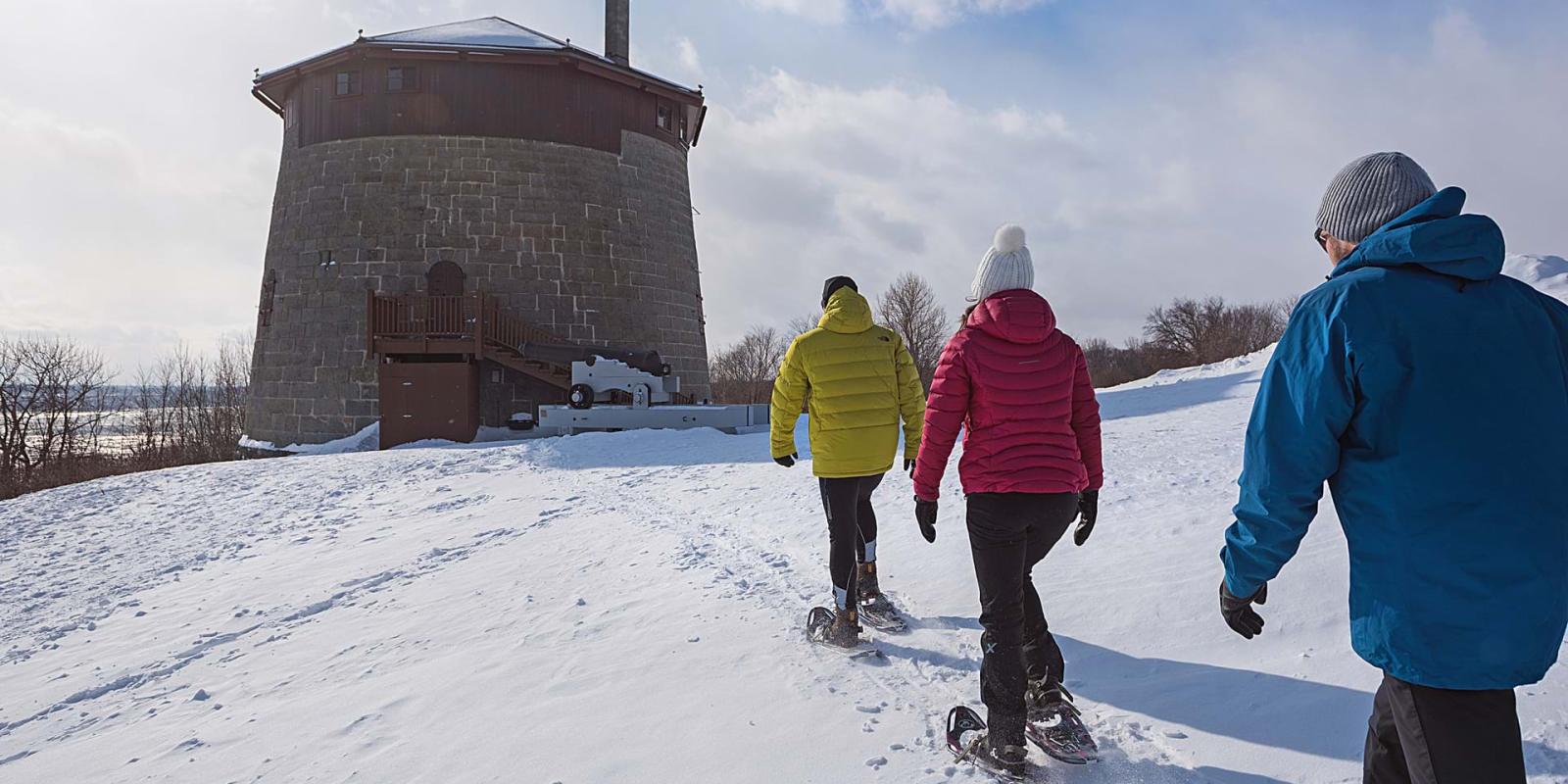Three friends are snowshoeing on the snow-capped Plains of Abraham and heading towards the historic Martello Tower.