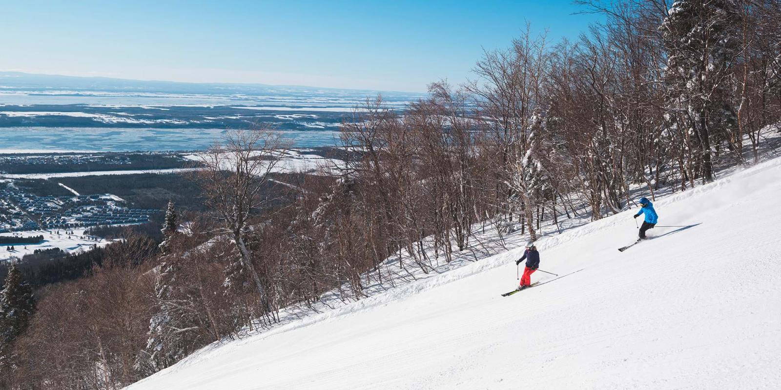 Two skiers go downhill skiing at Mont-Sainte-Anne station with a view of the St. Lawrence River.