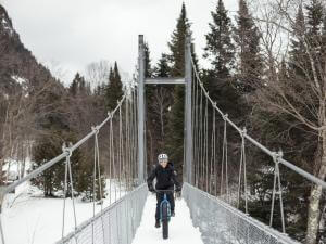 A cyclist rides a fatbike and crosses a snow-covered footbridge in the Vallée Bras-du-Nord.