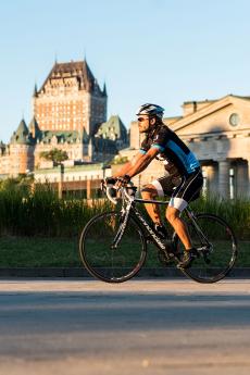 Un cycliste à vélo roule sur une piste cyclable dans le Vieux-Port de Québec avec la Château Frontenac en arrière-plan.
