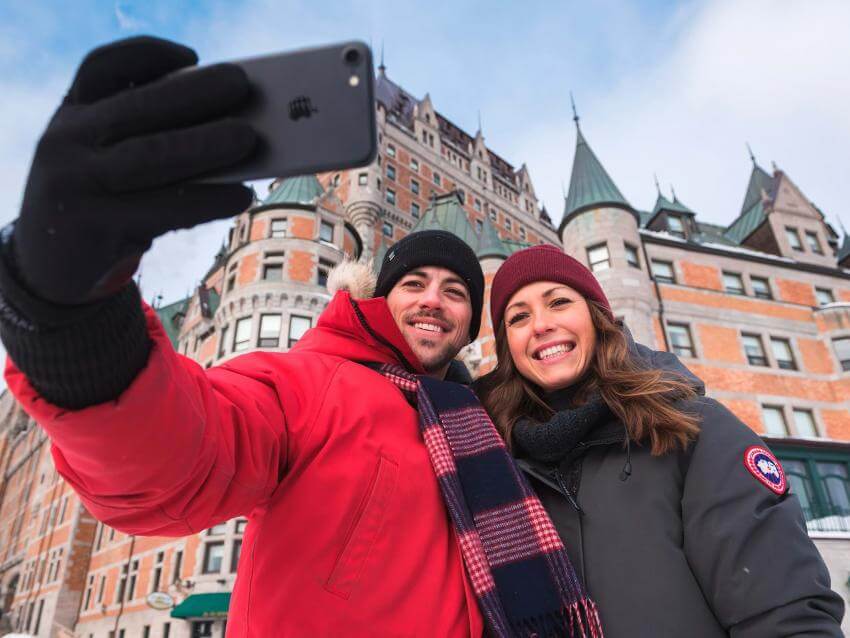 A smiling couple takes a picture of themselves in front of the Château Frontenac, in winter.