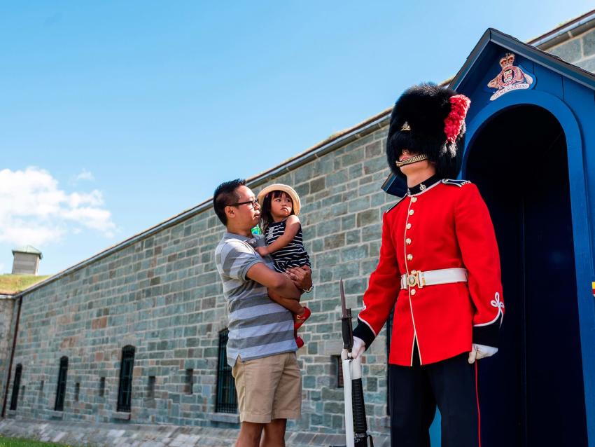 Un père et sa fillette regardent un garde vêtu de l'uniforme traditionnel devant la porte d'entrée de la Citadelle de Québec.
