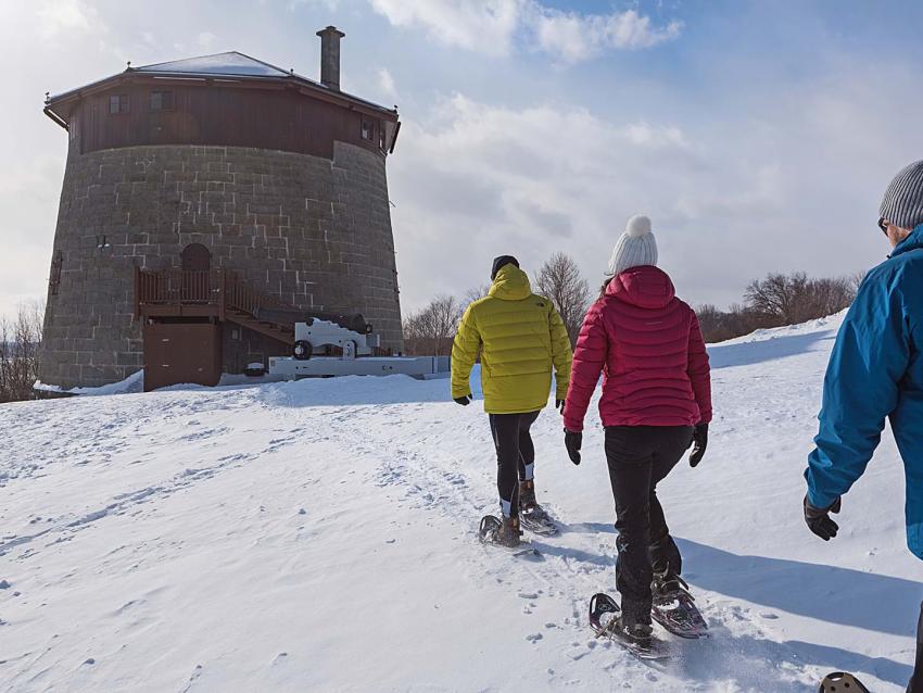 Three friends are snowshoeing on the snow-capped Plains of Abraham and heading towards the historic Martello Tower.