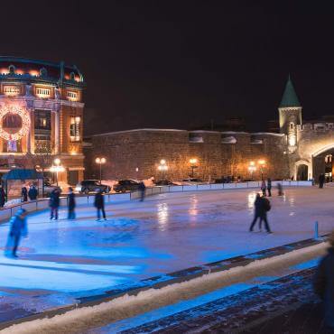 People on the outdoor ice rink at Place D'Youville, near the Capitol, the fortifications and the illuminated Saint-Jean gate.
