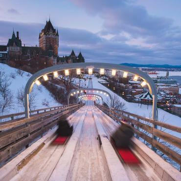 Two people slide on a tobogan at the Glissades de la Terrasse Dufferin, adjacent to the Fairmont Le Château Frontenac.