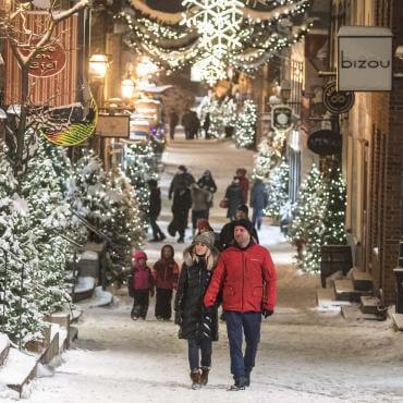  Several people walk in the evening on rue du Petit-Champlain, covered with snow and decorated with many illuminated trees.