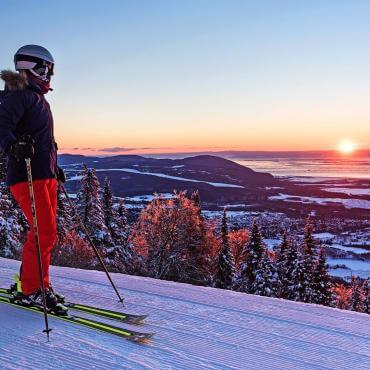 Une skieuse en ski alpin prend une pause et observe le coucher du soleil au sommet d'une montagne au Mont-Sainte-Anne.