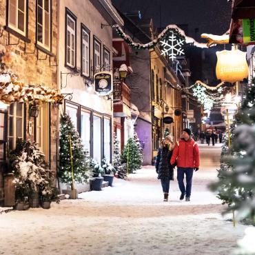 Un couple se promène en soirée durant le temps des fêtes sur la rue du Petit-Champlain enneigée et décorée de sapins illuminés. 